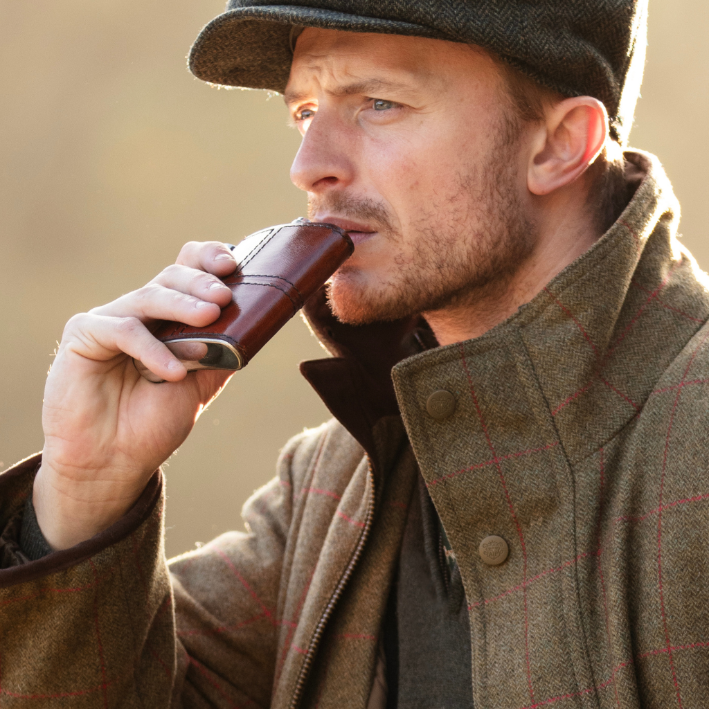 A man in a brown tweed cap and matching plaid jacket drinking from a brown leather hip flask outdoors.