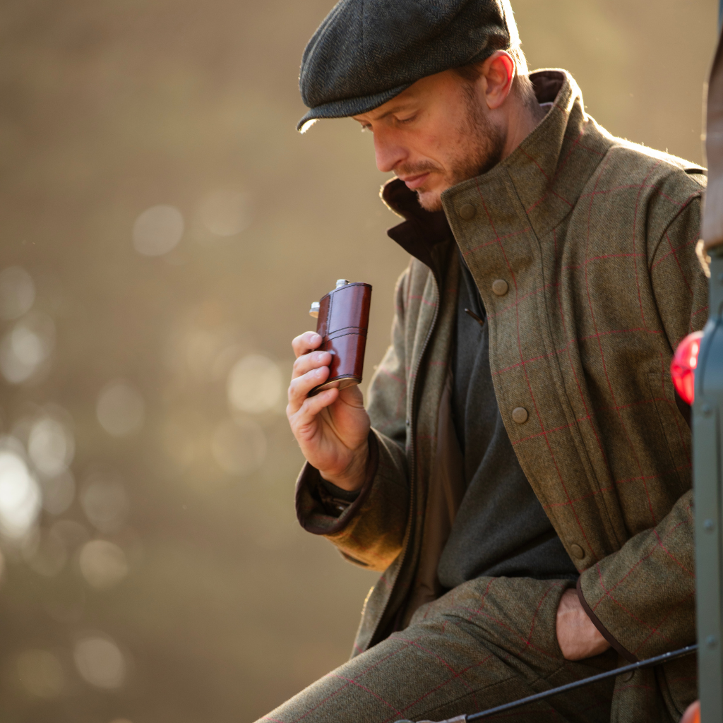 A man wearing a dark flat cap and a brown tweed overcoat with red windowpane plaid, holding a small brown leather hip flask. The background is a soft, warm, blurred bokeh of golden light.
