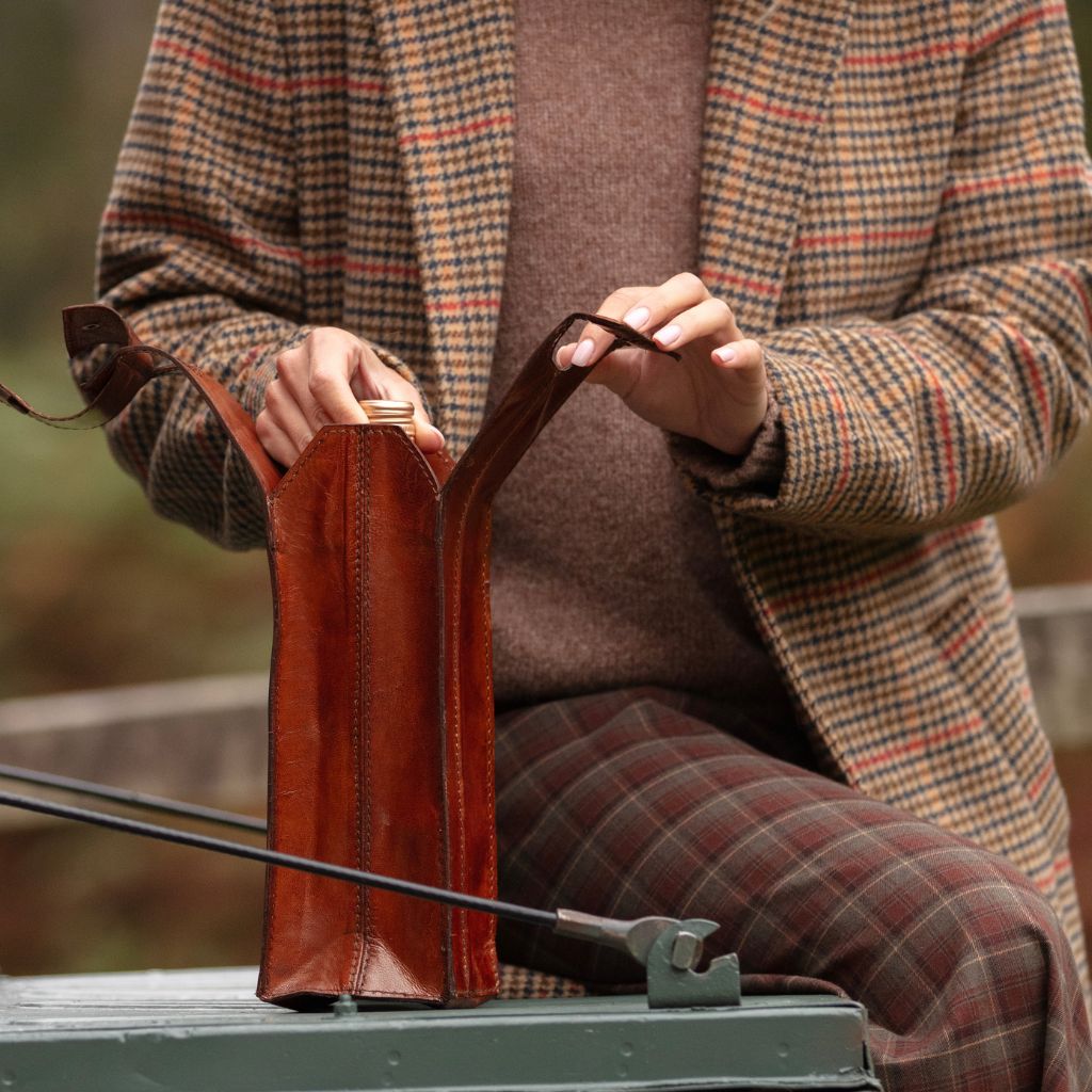 A person wearing a brown and beige plaid blazer and matching trousers holds a leather wine carrier and is sittiing on the tailgate of a Landrover