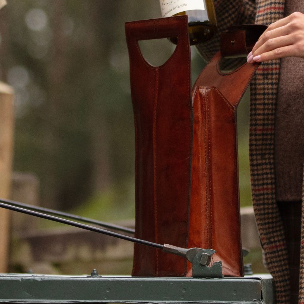 A close-up of a person pouring wine from a bottle into a leather wine carrier sitting on the tailgate of a landrover