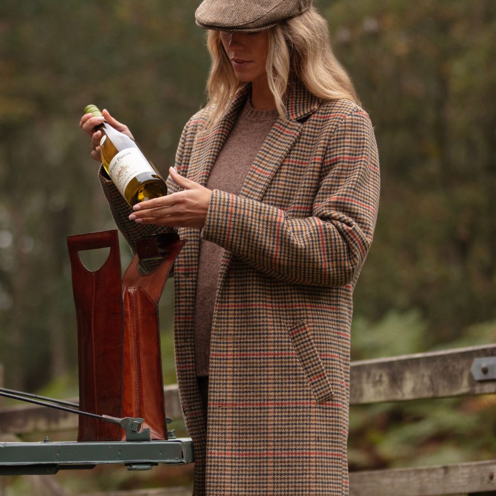 A woman wearing a brown plaid overcoat and matching flat cap stands outdoors near a wooden fence, holding a wine bottle and a brown leather wine holder.
