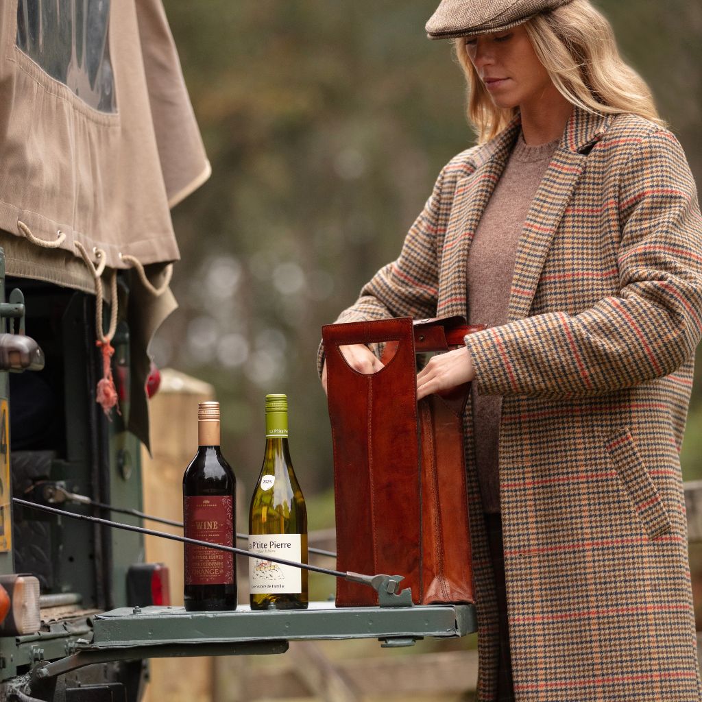 A woman in a brown tweed plaid coat and flat cap stands beside a vehicle, putting wine into a leather carrier next to two wine bottles on a landrover tail gate