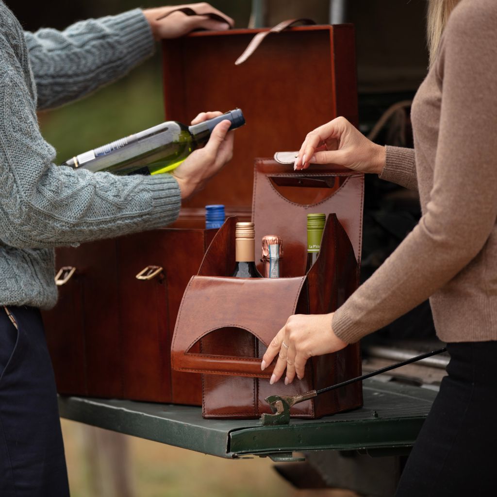 A brown leather wine bottle carrier for four bottles of wine h being handled by two people at a table with a leather wine trunk for six bottles in the background.