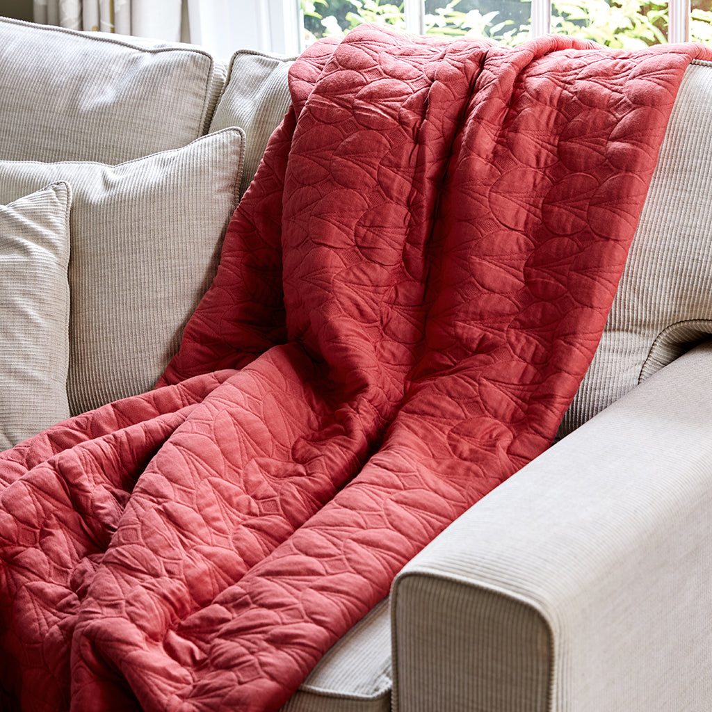 A coral-colored, tulip design quilted cotton bedspread draped over the back of a beige sofa, with natural light illuminating the fabric.