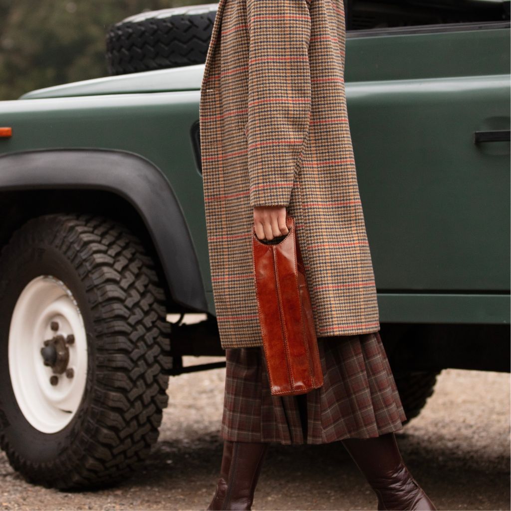 A person wearing a long brown and beige plaid coat, a matching plaid skirt, and dark brown boots stands beside a green off-road vehicle, holding a brown leather single wine carrier.