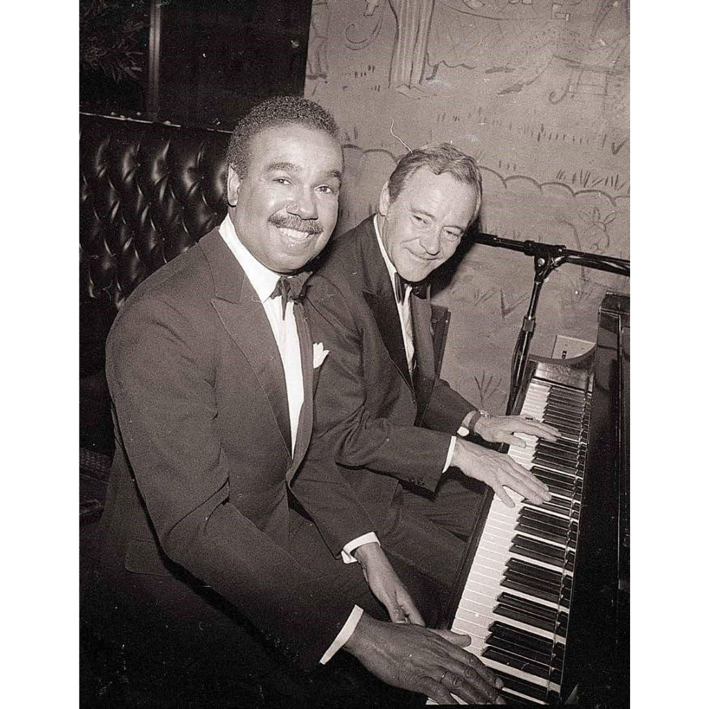 Two men in suits sitting at a piano, posing for a photo.