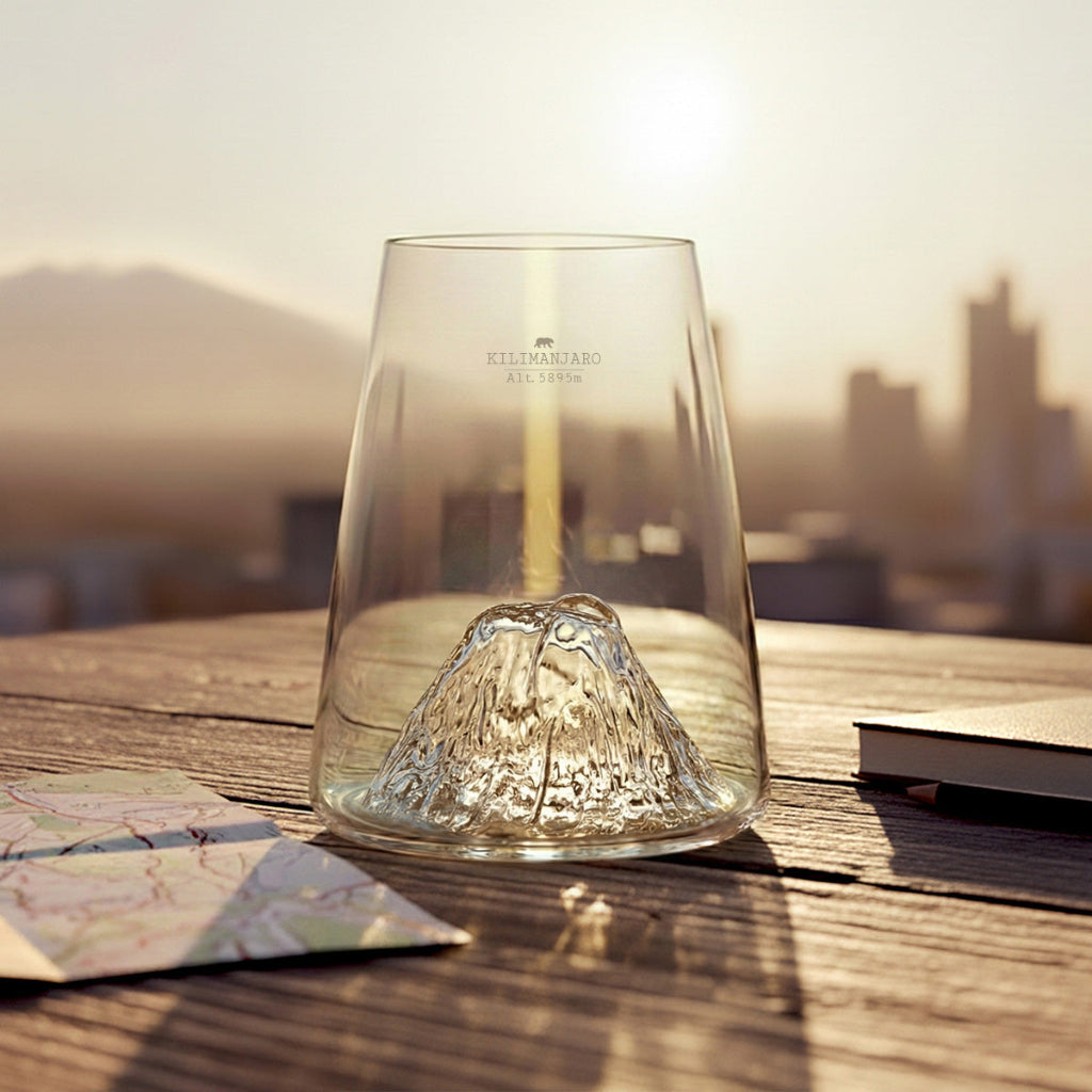 A clear glass with a textured mountain design inside, set on a wooden table with a blurred cityscape and mountains in the background at sunset.