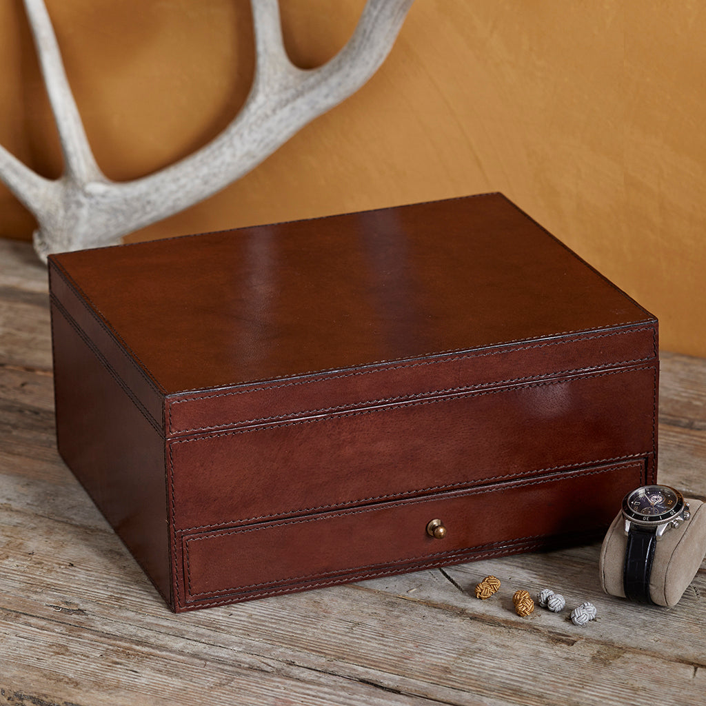 A  leather watch box with a single drawer styled on a rustic wooden surface with a watch.