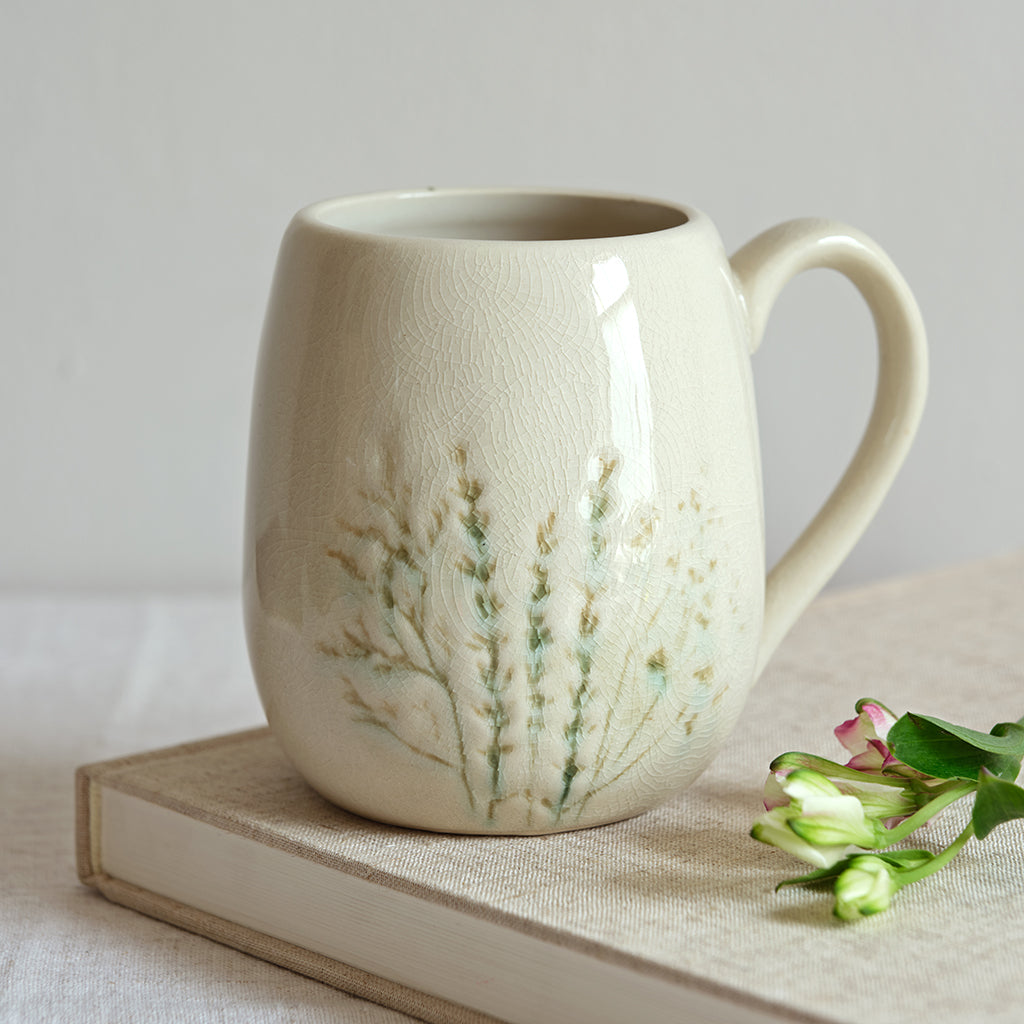 White ceramic mug with floral design on a light surface with a book and flowers.
