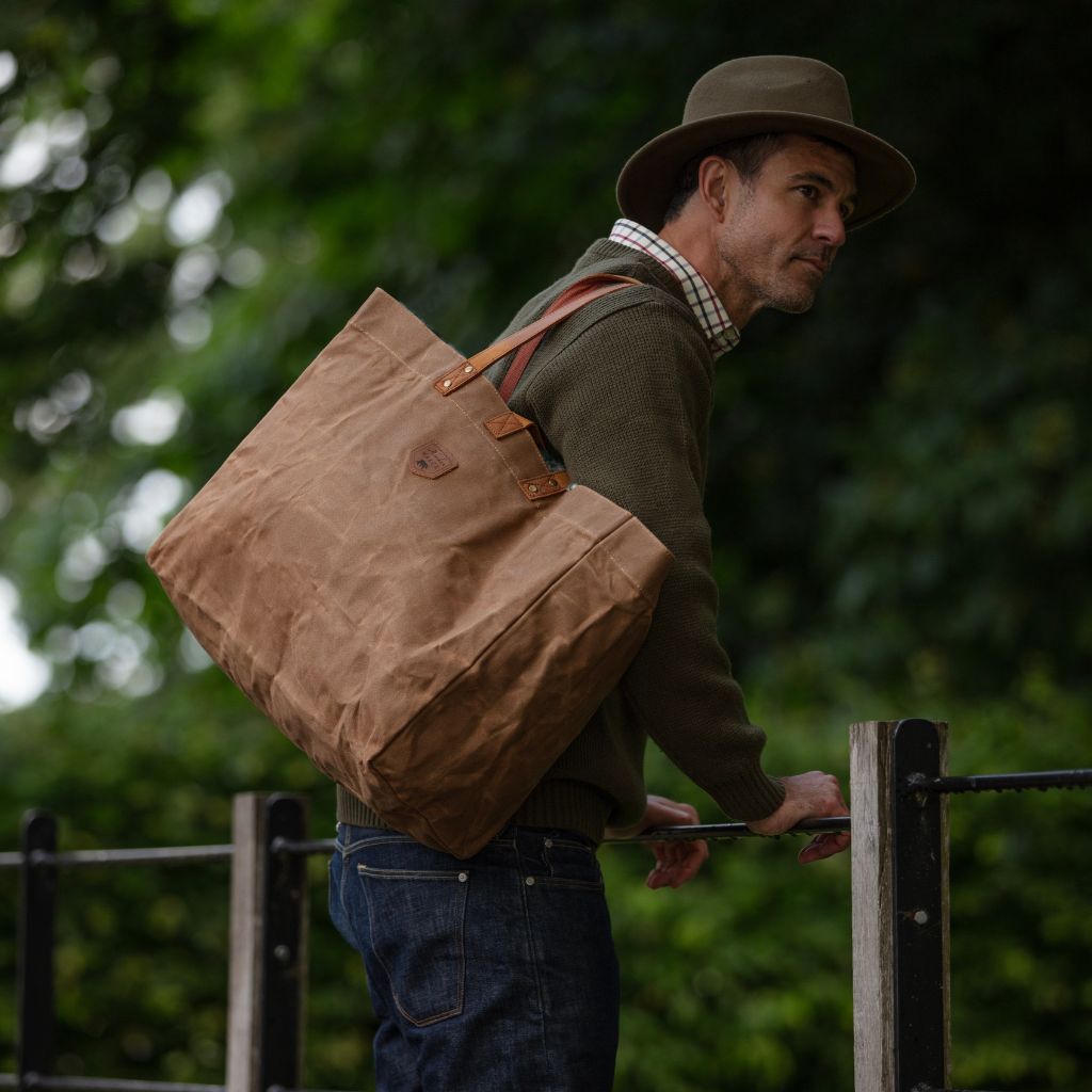 Man with a brown waxed canvas bag over his shoulder, standing outdoors with greenery in the background.
