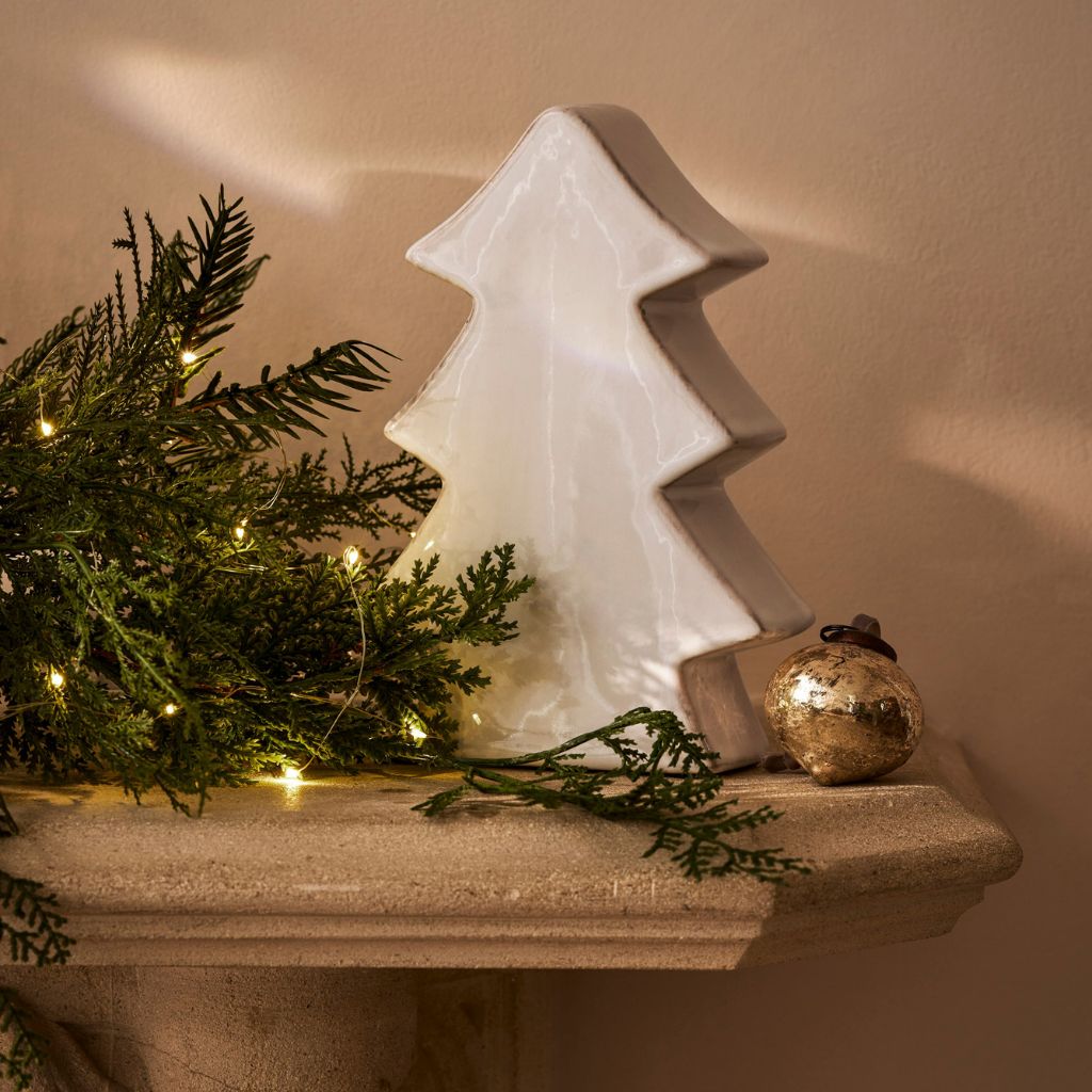 Decorative Christmas scene with a white ceramic tree, greenery, and a gold ornament on a wooden surface.