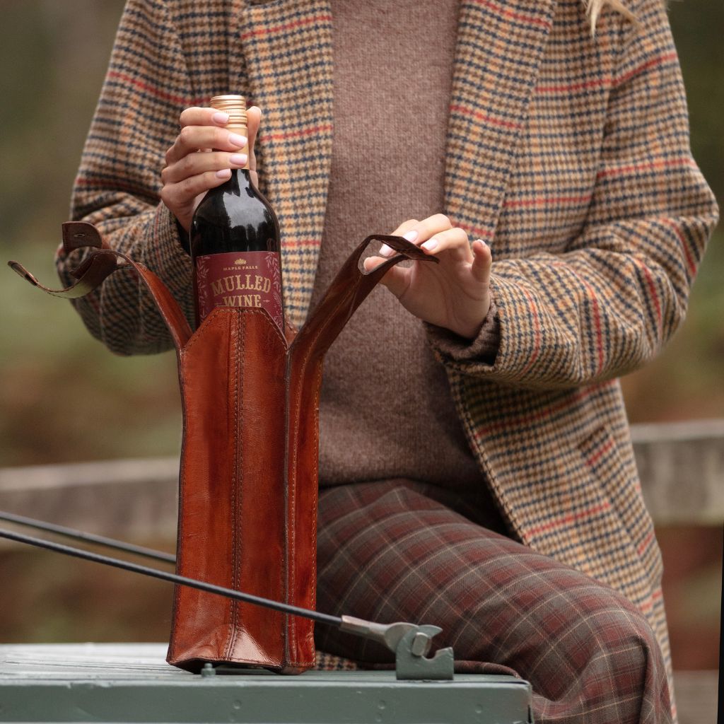A person wearing a brown and beige plaid coat and matching plaid trousers holds a dark wine bottle in a brown leather wine carrier.