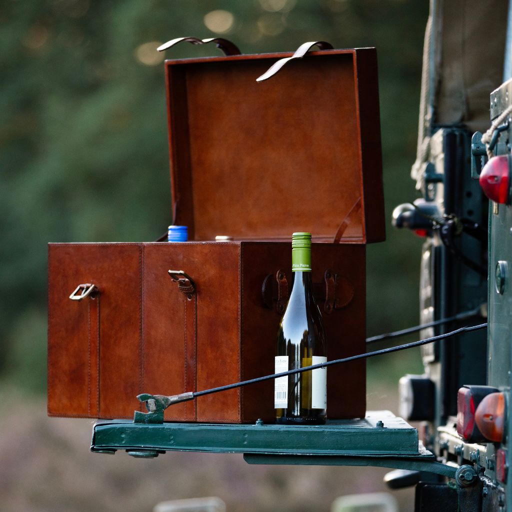 A brown leather wine trunk with an open lid, mounted on the back of a green vehicle. A wine bottle with a green cap is placed inside the case, and the background shows a blurred outdoor setting.