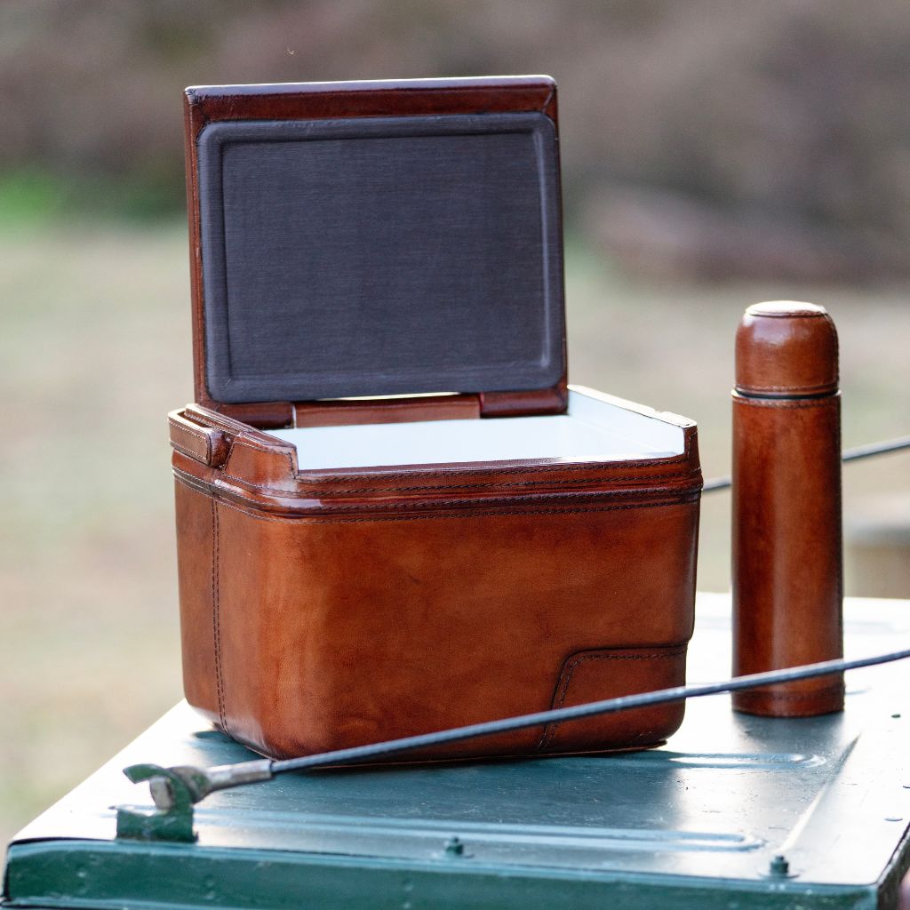 A brown leather cooler case with its lid open, shown outdoors next to a matching brown bottle on the tailgate of a landrover