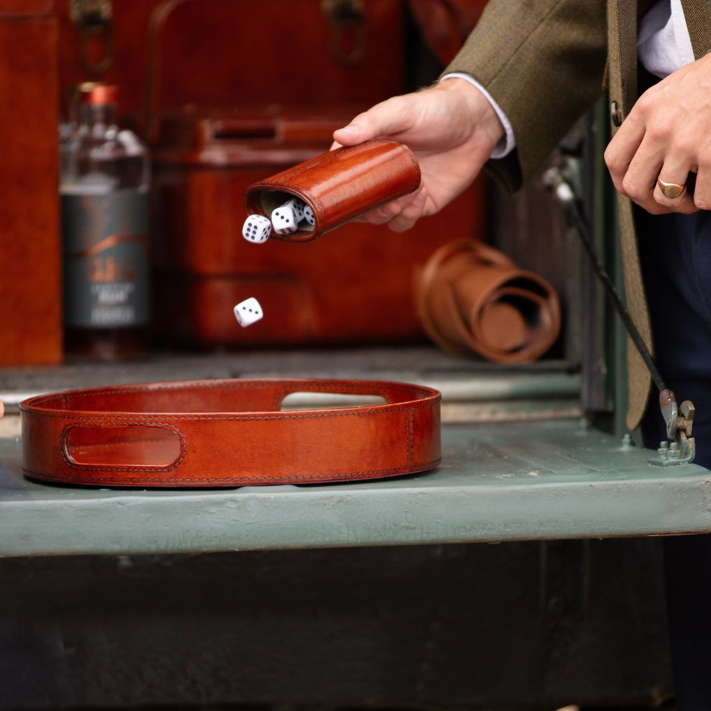 A person in a brown jacket holds a small brown leather dice cup shaker while rolling white dice, with a matching brown leather tray resting on a table in a styled indoor setting.