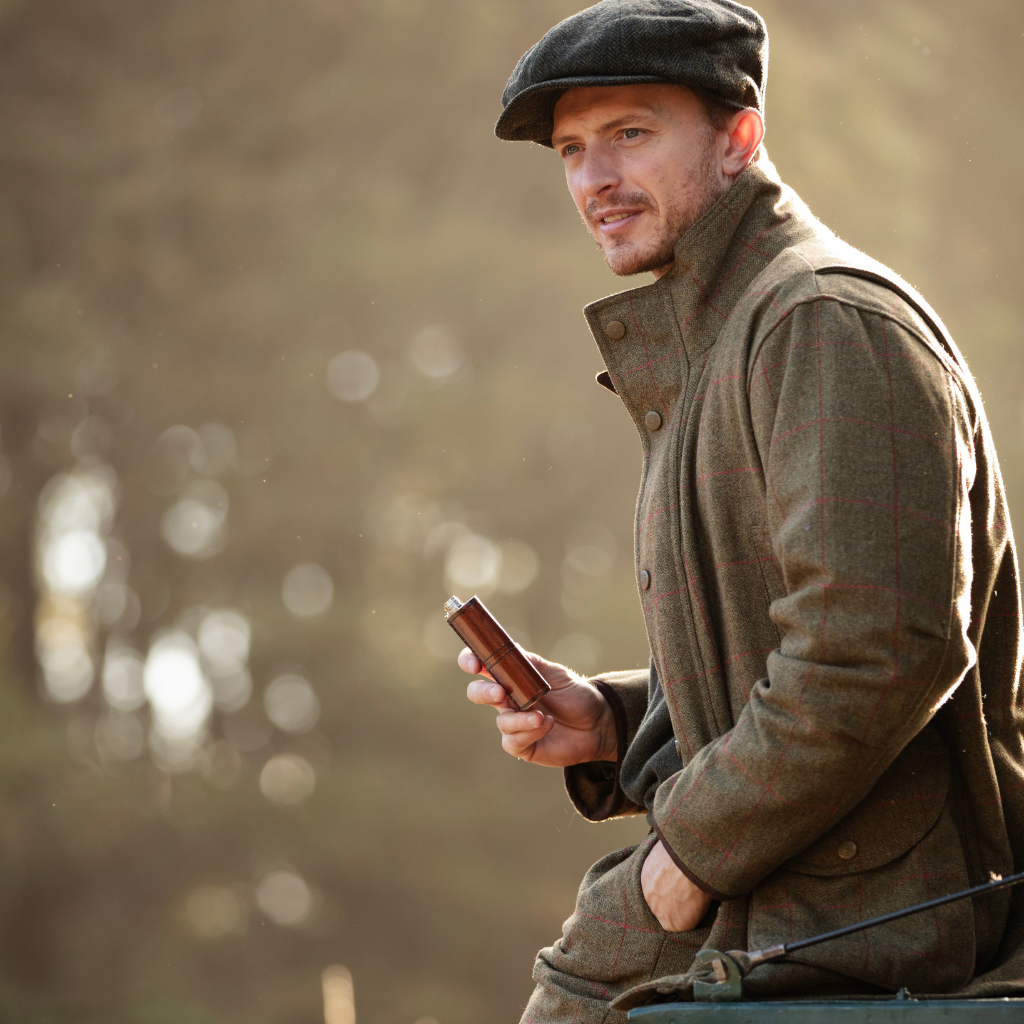 A man wearing a dark flat cap and a brown tweed hunting jacket, holding a brown leather hip flask, set against a warm, blurred outdoor background.