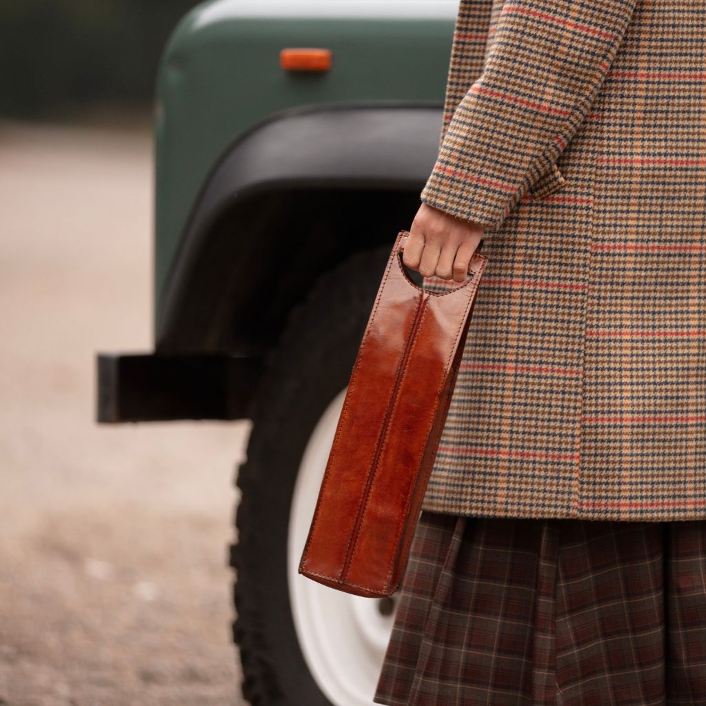 A person in a brown and beige plaid coat and matching skirt holds a brown leather single wine carrier standing beside a vintage green vehicle outdoors.