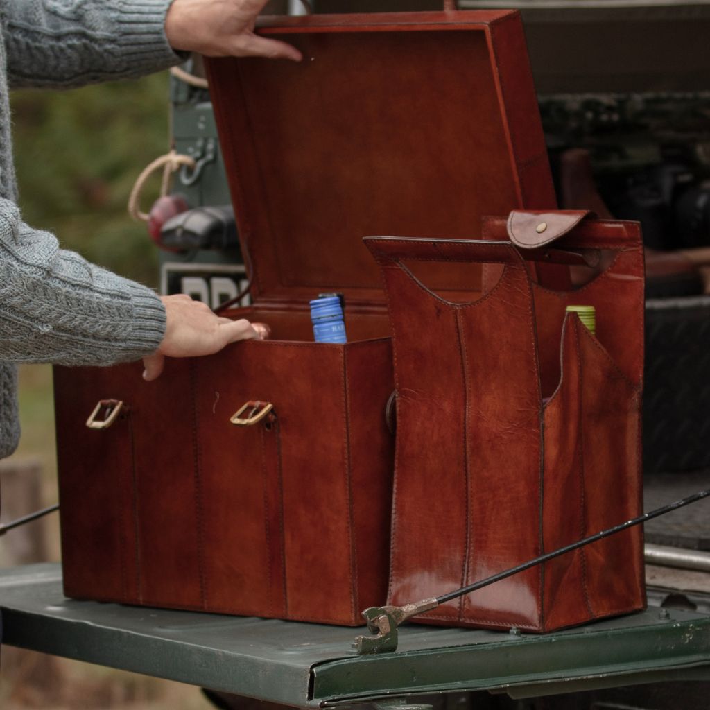 A person wearing a gray knit sweater opens a wine trunk on a landrover tailgate, with a matching smaller  leather wine carrier for four bottles. 