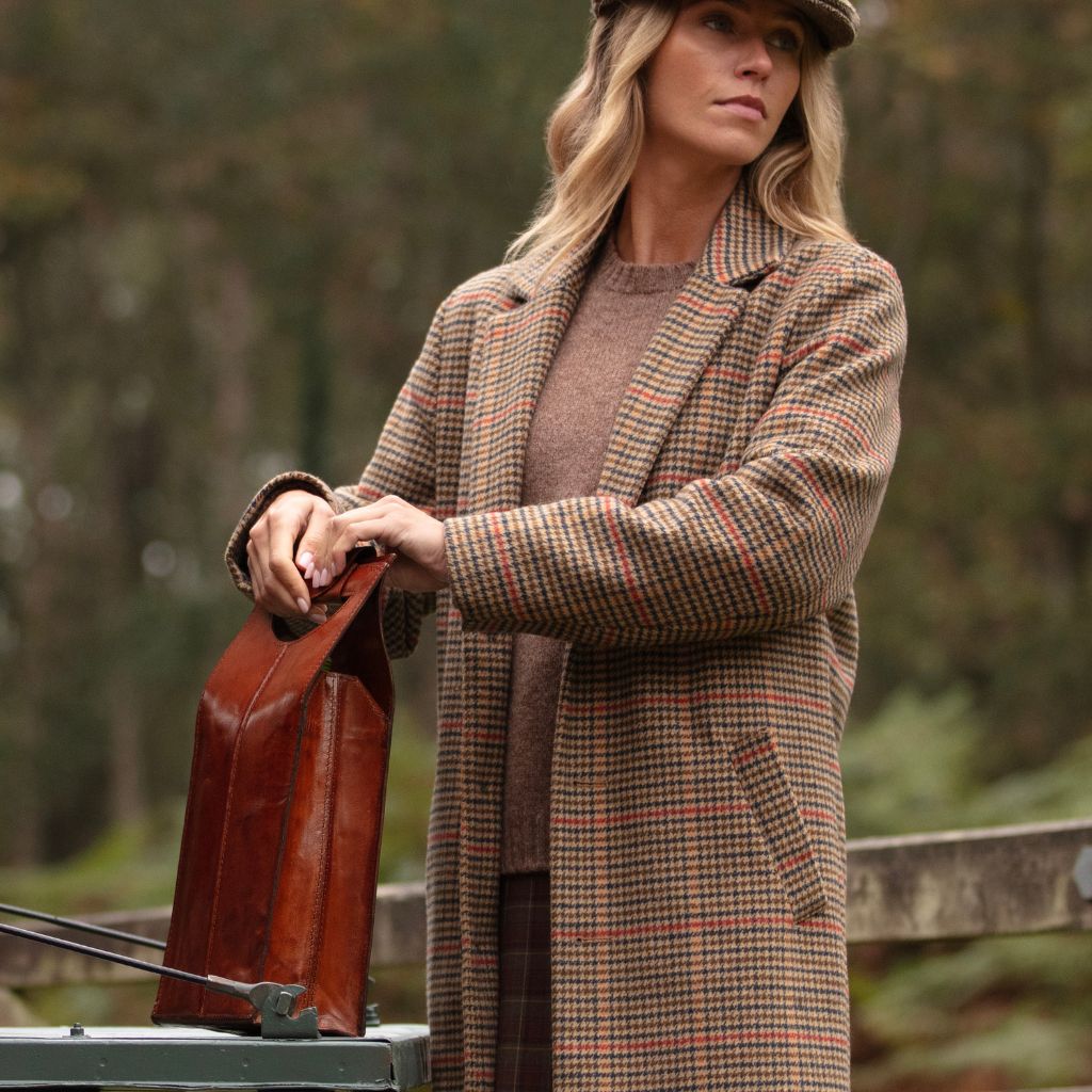 A woman wearing a brown and beige plaid overcoat and a brown hat stands outdoors holding a double leather wine carrier with a blurred forest background.