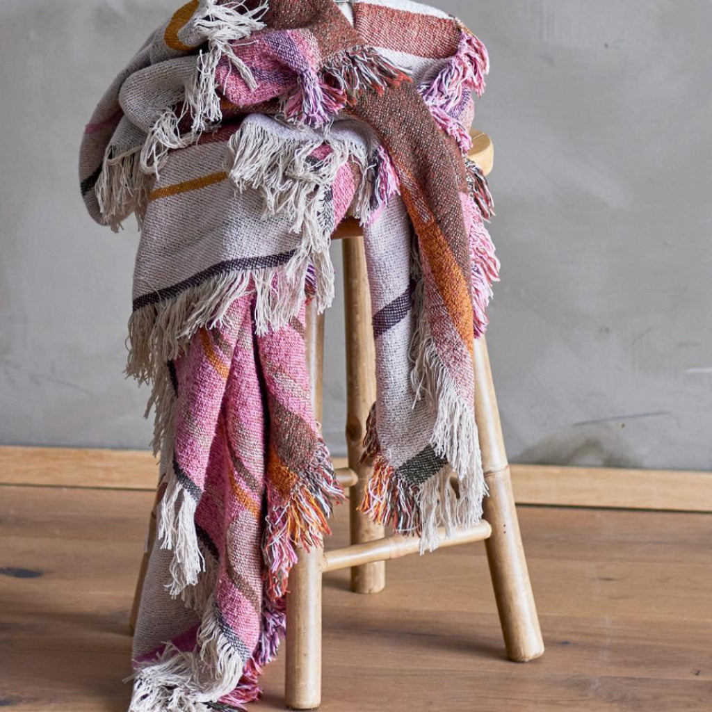 Woven blanket with fringes draped over a wooden stool against a grey wall.