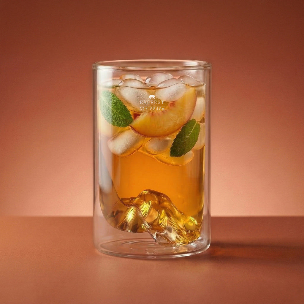 A clear double-walled glass tumbler with a sculpted mountain-shaped at the bottom, filled with an amber drink, ice cubes, a peach slice, and mint leaves, set against a warm brown gradient background.