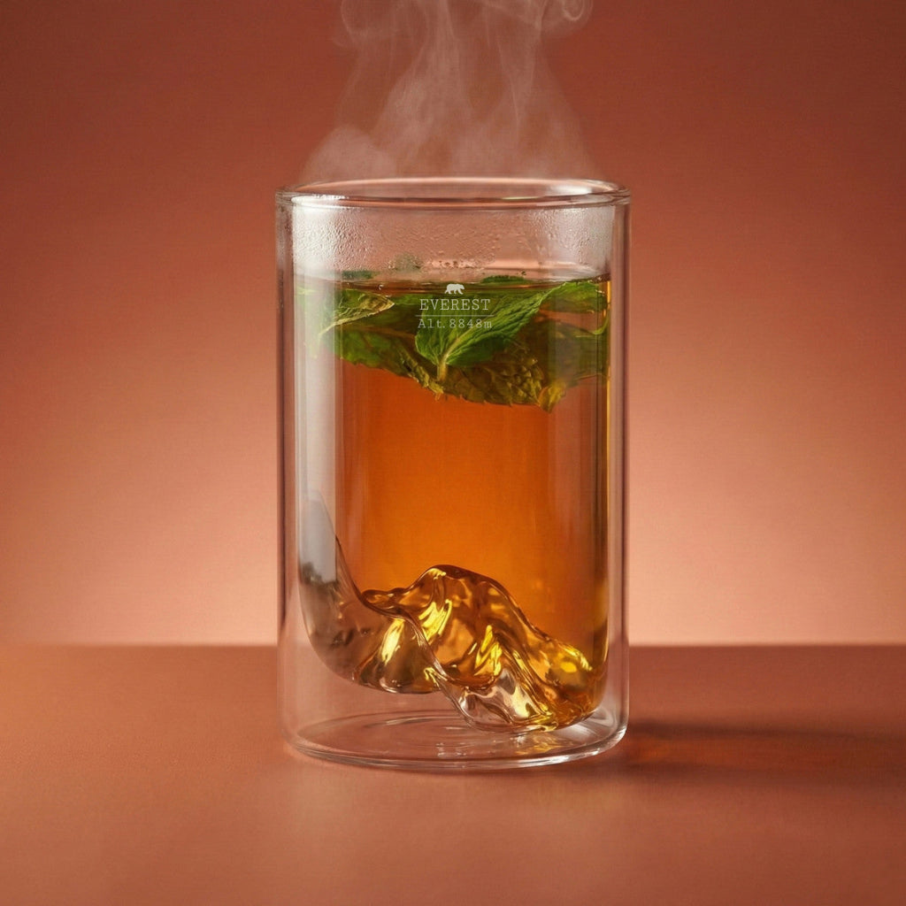 A double-walled clear glass mug with a sculpted mountain-shaped base, filled with amber tea, mint leaves, set against a warm peach-brown gradient background.