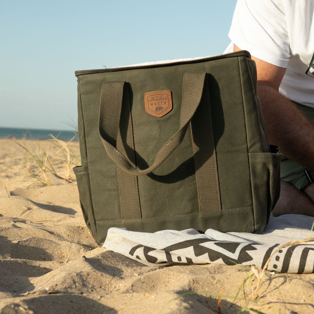 An olive green canvas cool bag with dual webbing handles and a brown leather patch logo, sitting on a sandy beach next to a person wearing a white t-shirt.