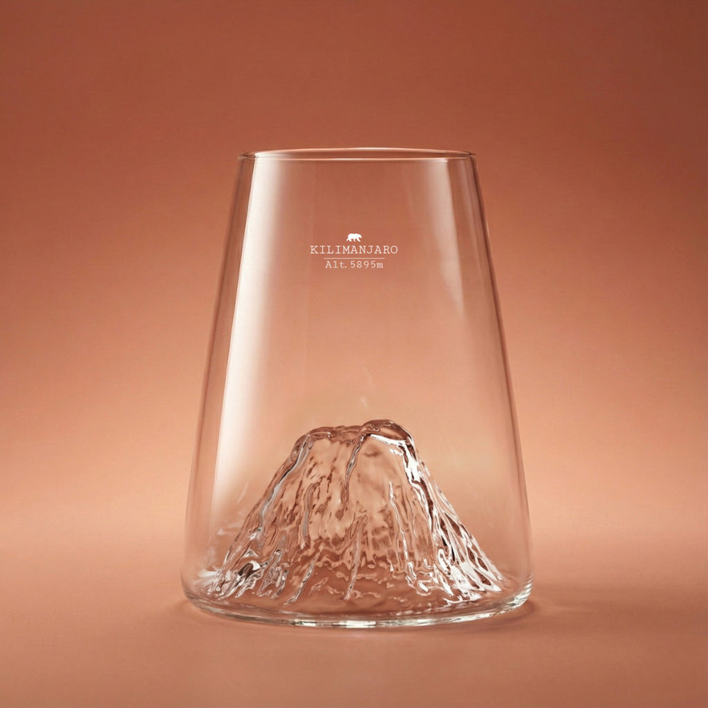 A clear glass tumbler with a sculpted mountain design at the base set against a warm terracotta background.