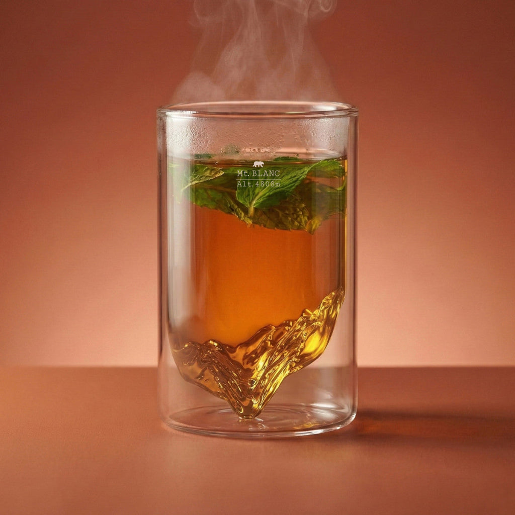 A clear double-walled glass mug with a mountain-shaped bottom, filled with amber tea and a mint leaf, set against a warm terracotta background with steam rising.